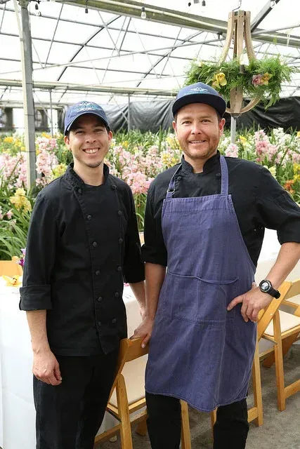 Two chefs in black uniforms and aprons standing in a greenhouse with hanging flowers, smiling at the camera