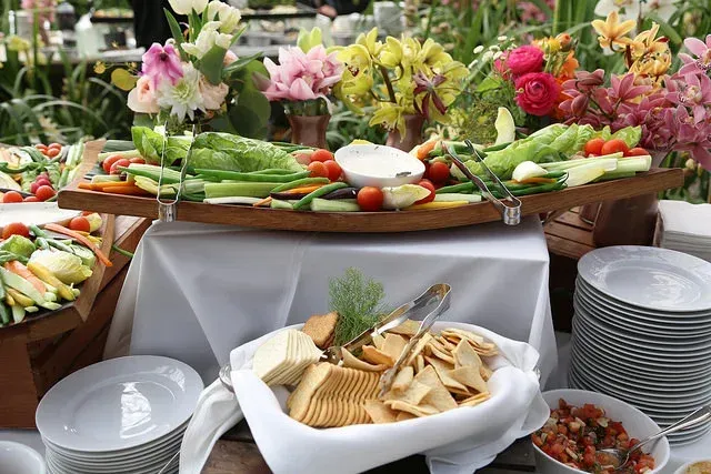 Buffet table with assorted salads, flowers, dips, and stacks of white plates at an outdoor event