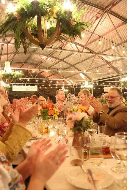 Guests clapping at a candlelit banquet table under hanging greenery in a warmly lit event hall