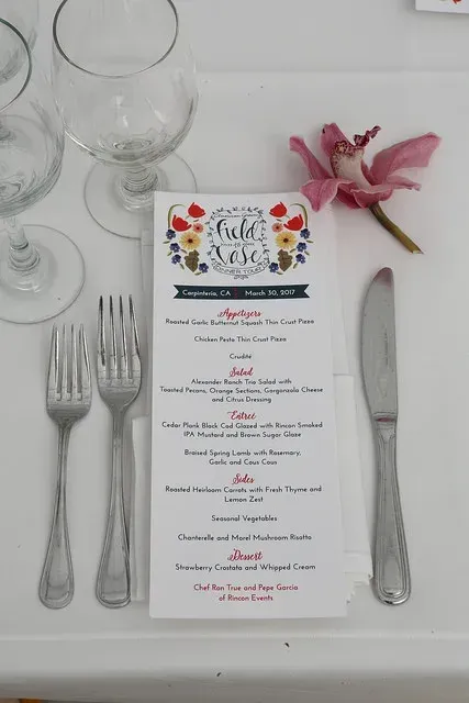 Festive floral place setting with menu, silverware, wine glasses, knife, and pink flower on a white tablecloth