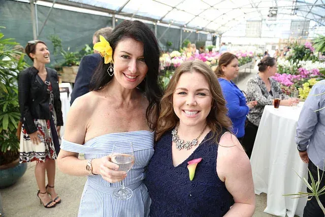 Two smiling women posing at a flower-filled indoor event, one holding a drink