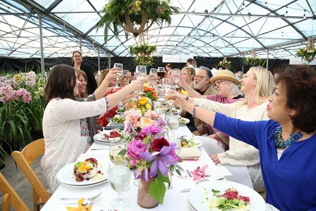 Guests toast at a long table in a bright glass greenhouse filled with flowers and plants.