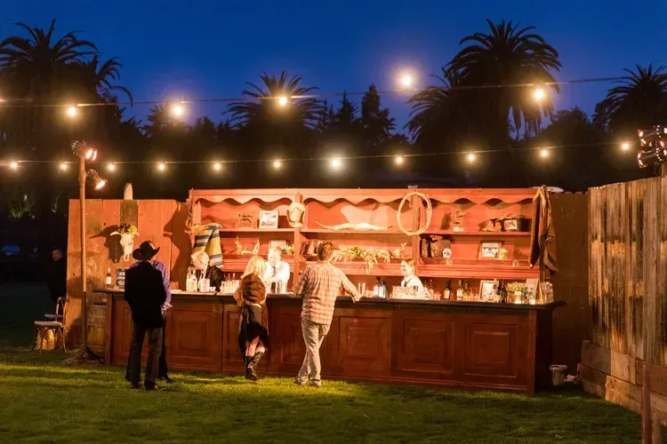 Outdoor bar at dusk with warm string lights and several people standing at the counter in a backyard garden.