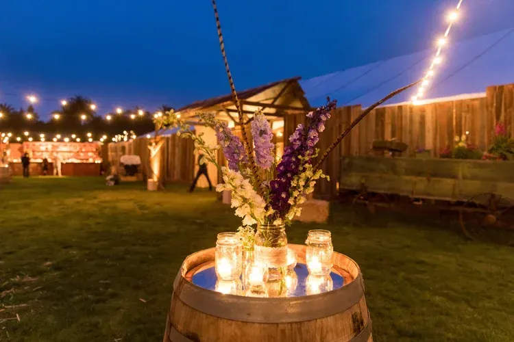 Outdoor evening wedding reception with lit barrel centerpiece and string lights over a grassy lawn