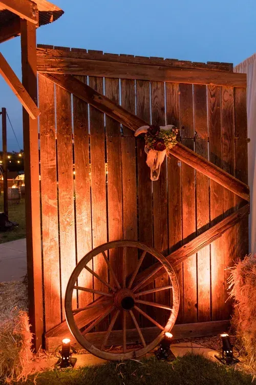 Rustic wooden gate lit by warm lanterns, with a wagon wheel and diagonal beam detail at dusk