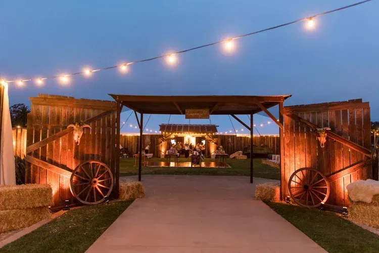 Rustic outdoor wedding setup at dusk with string lights, wooden archway, and candlelit tables in the distance
