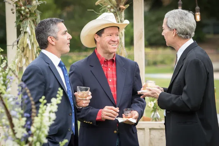 Three men in suits and a cowboy hat chatting and holding drinks outdoors at a garden event