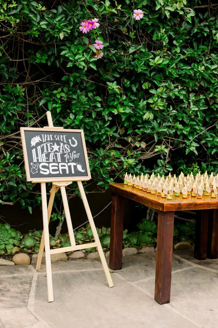 Chalkboard sign beside a wooden table of small bottles against a leafy green wall.