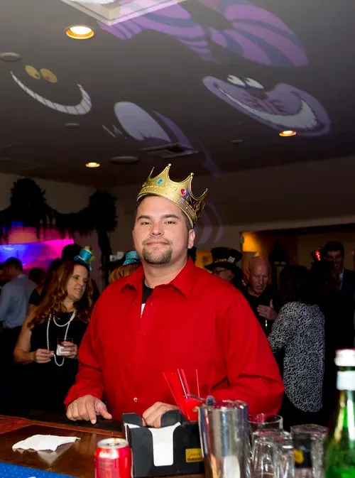 Man in a red shirt wearing a gold crown at a party bar, standing behind drinks
