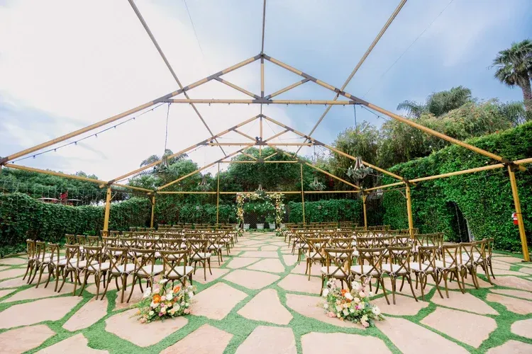 Outdoor event setup with rows of chairs under a white canopy, surrounded by greenery and stone paving