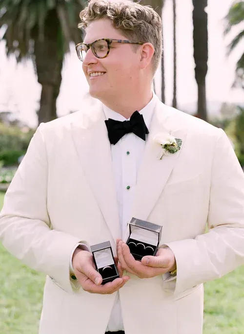 Man in a white tuxedo holding two ring boxes, smiling outdoors.
