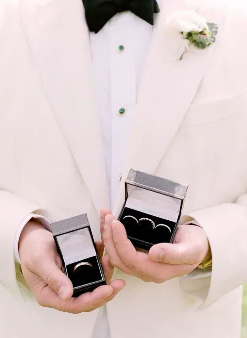 Person in a white tuxedo holds two open ring boxes with black interiors.