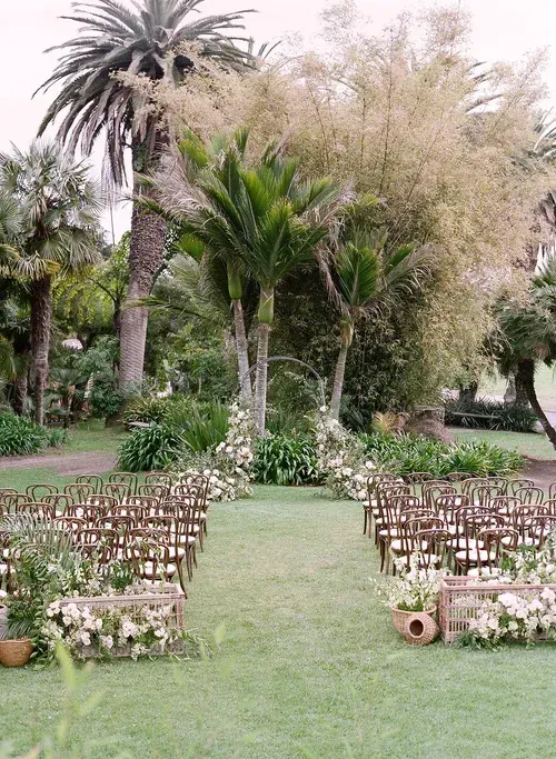 Rows of white chairs on a grassy lawn in a garden with tall palm trees and a floral arch