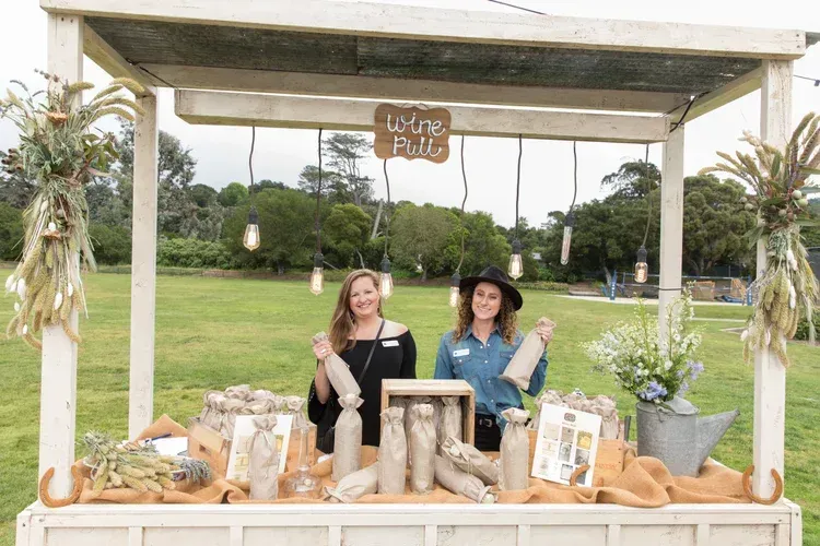 Two people posing at an outdoor booth with plants, logs, and a sign reading “Love Bloom.”