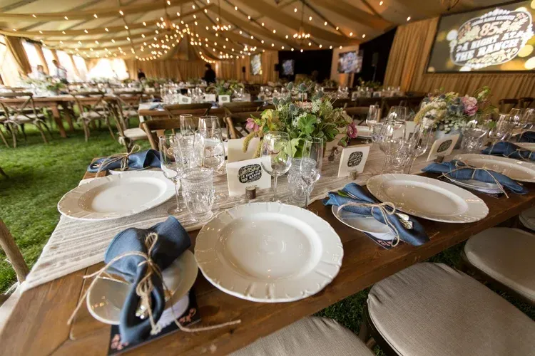 Decorated wedding reception table with blue napkins, plates, glassware, and warm string lights in a tent