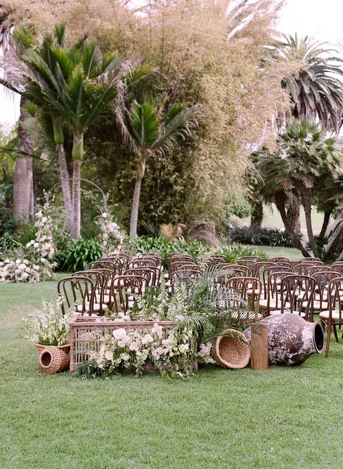 Outdoor garden seating with wooden chairs, floral arrangements, and woven decor on a green lawn beneath palm trees