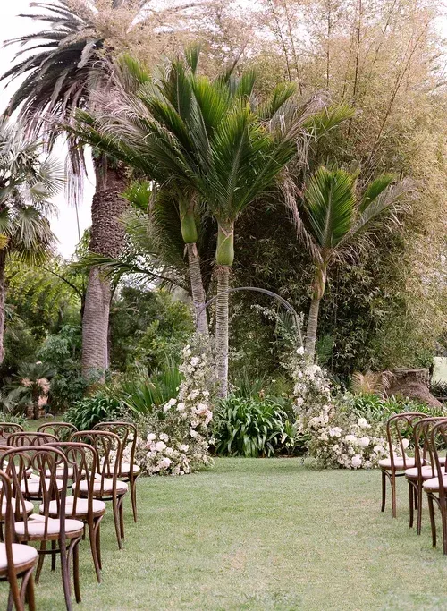 Outdoor garden ceremony aisle lined with wooden chairs, surrounded by tall palm trees and lush greenery