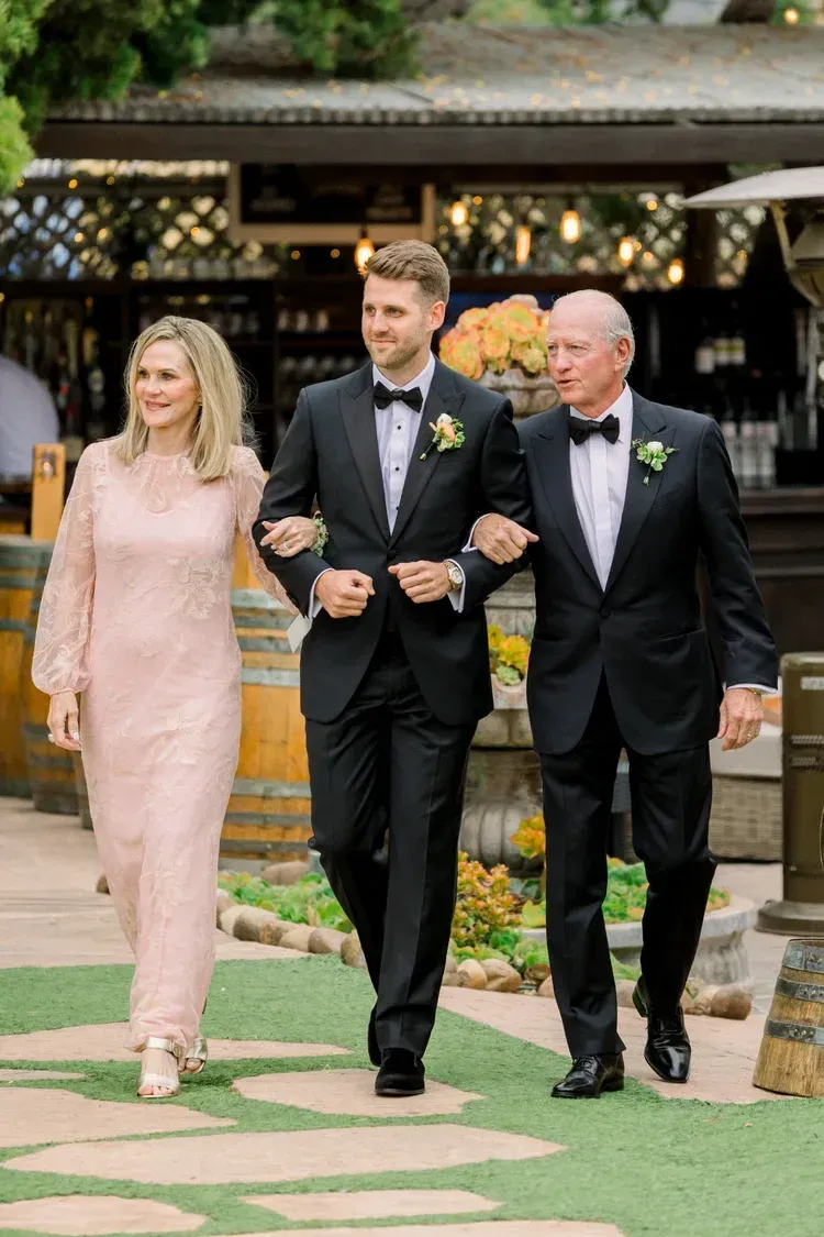 Three people in formal wear walking arm in arm outdoors at a wedding venue