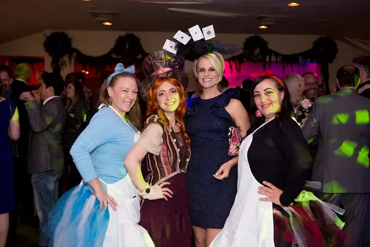 Four women in costumes pose with hands on hips at a lively party dance floor.
