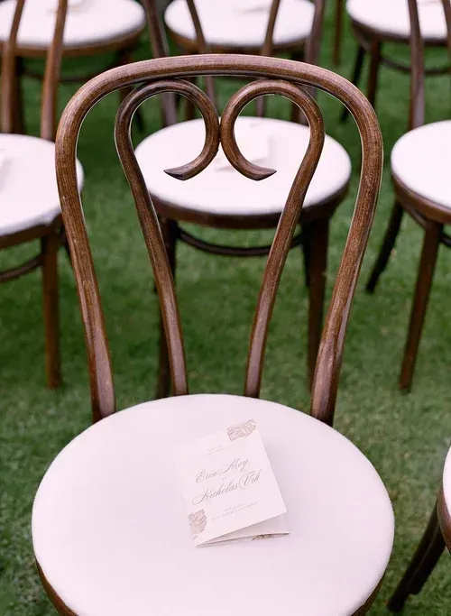 Rows of dark wooden chairs with white seat covers and folded place cards on a green carpet.