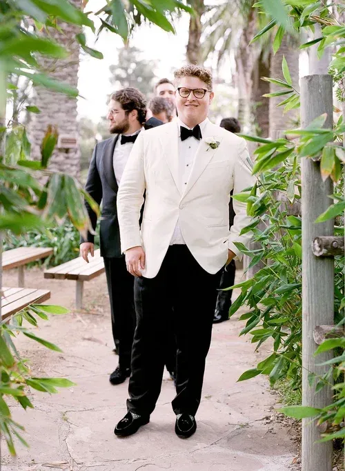 Man in white tuxedo stands in a garden path, with another man in a dark suit behind him.