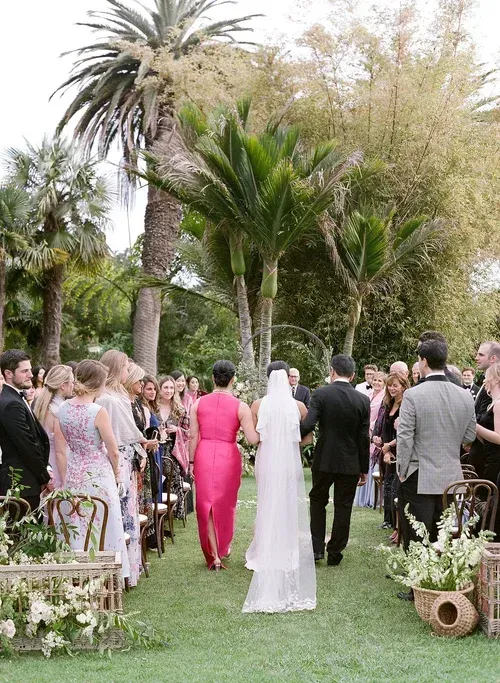 Wedding guests walk down a palm-lined garden aisle behind a bride in a white veil and a woman in a bright pink dress