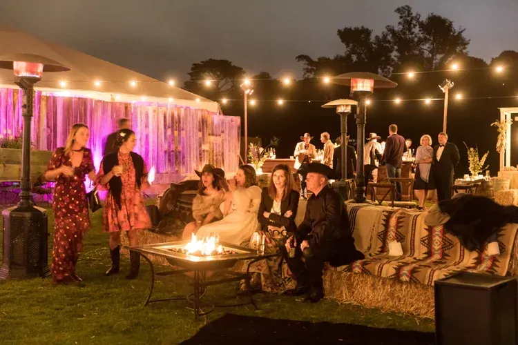 Outdoor evening party with string lights, guests seated on hay bales, and a fire pit glowing in the center