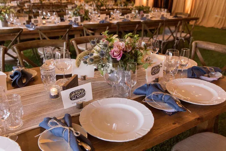 Elegant outdoor dining table set with glassware, blue napkins, candles, and floral centerpiece at dusk