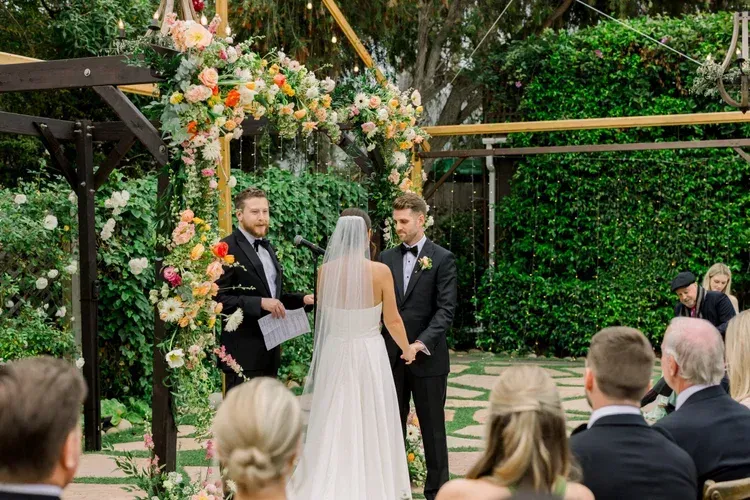 Wedding ceremony outdoors under floral arch, bride facing officiant and groom before seated guests