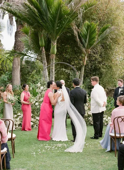 Wedding ceremony outdoors under palm trees, with couple exchanging vows and guests standing nearby.