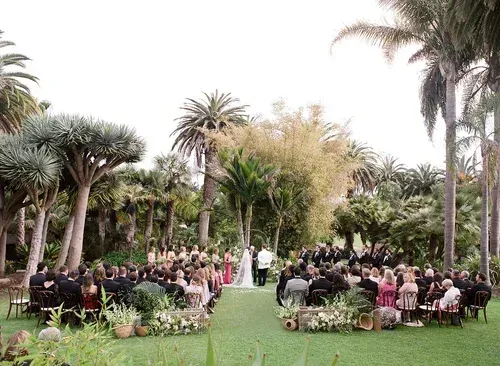 Outdoor wedding ceremony in a lush garden, with guests seated in rows facing an altar aisle.