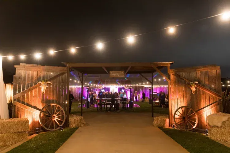 Outdoor event venue entrance with string lights, wooden gates, and a lit tent in the background