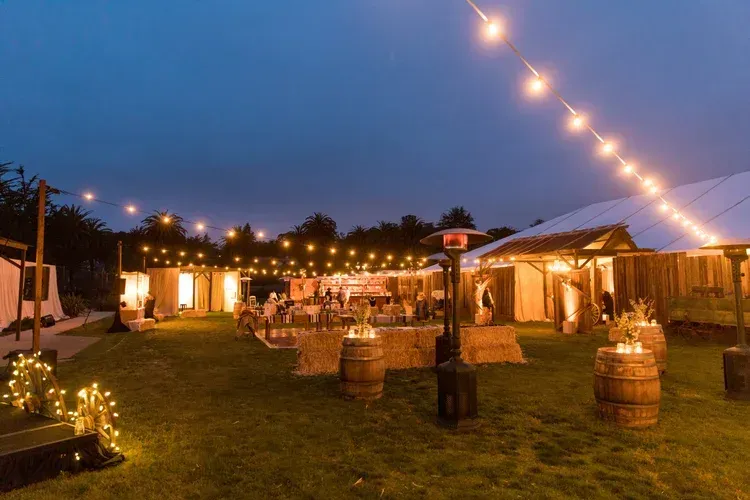 Twilight outdoor reception with string lights, tables, and barrels on a grassy lawn