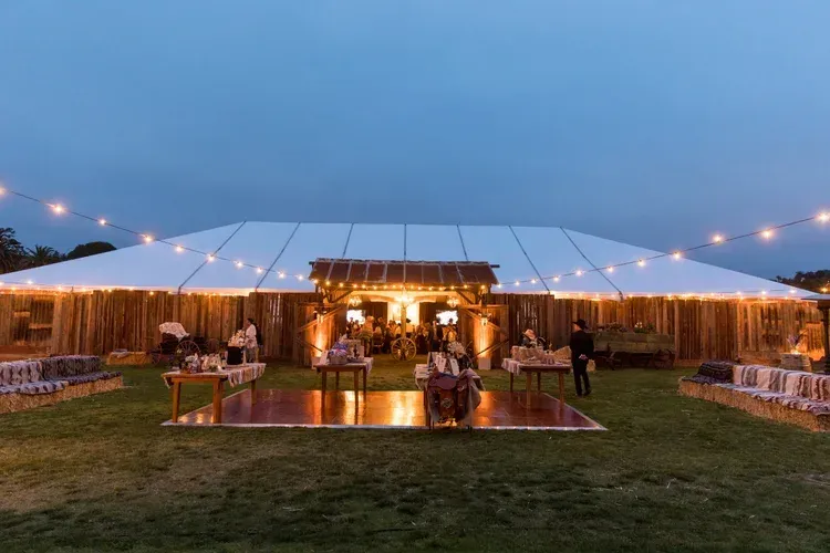 Lit white event tent at dusk with guests on a lawn and string lights glowing inside and around it