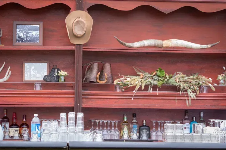 Red wooden bar shelves with bottles, glassware, hats, framed photos, and animal skull decor
