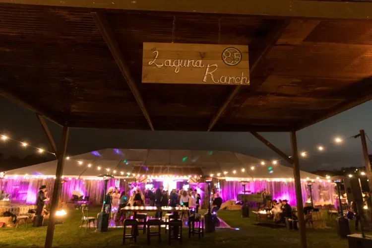 Outdoor wedding reception under a lit pavilion at dusk, with tables and guests on a lawn.