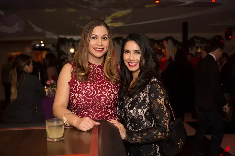 Two women posing at a dimly lit event bar, one in a red dress and one in a black lace top