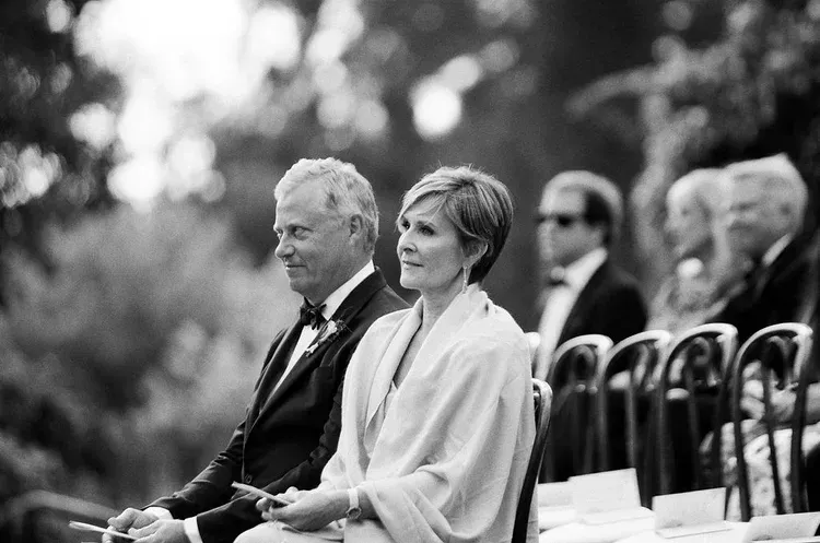 Seated guests at an outdoor ceremony, including a woman in light clothing in the foreground.