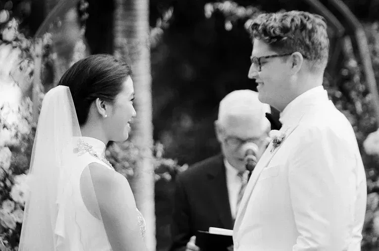 Bride and groom face each other during an outdoor wedding ceremony, with an officiant behind them.