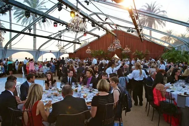 Crowded indoor reception under a glass roof, with guests seated at round tables and mingling near a red wall.