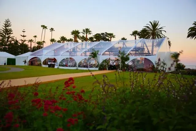 White event tent at dusk in a garden, with palm trees and red flowers in the foreground.