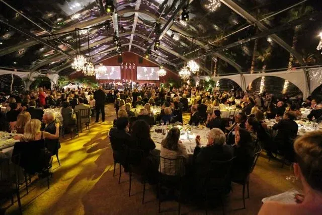 Crowded banquet under a lit tent with many seated guests and a stage at the far end