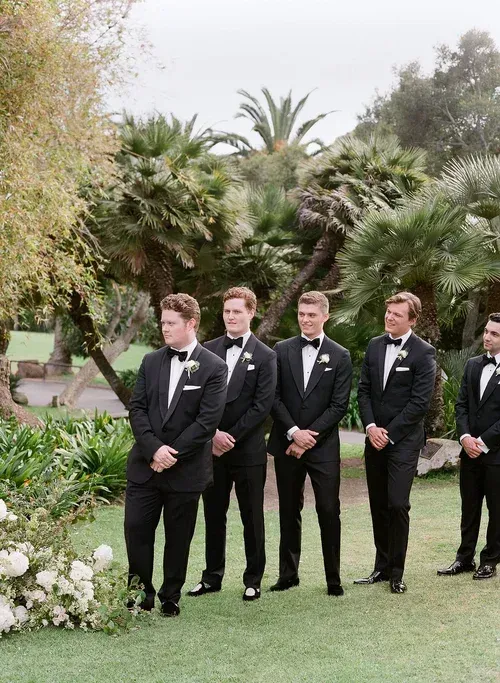 Groomsmen in black tuxedos standing on a garden lawn during an outdoor wedding ceremony