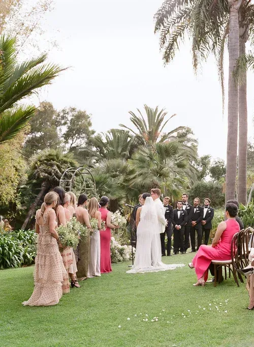 Outdoor wedding ceremony on a lawn, with bride, groom, bridesmaids, and guests surrounded by palm trees.