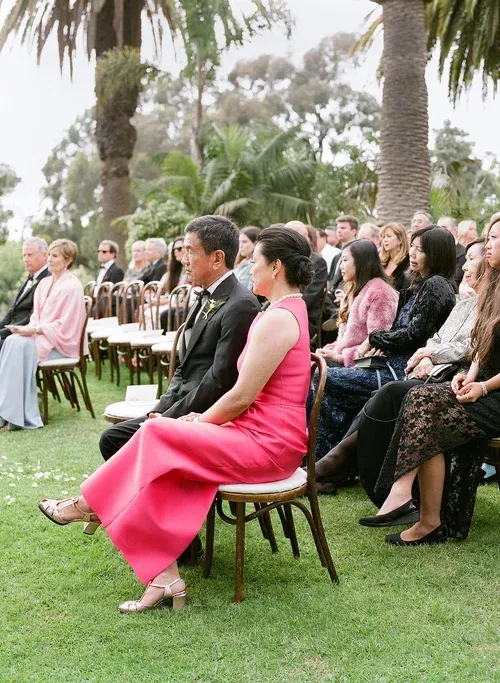 Guests seated outdoors at a formal ceremony, with a woman in a bright pink dress in the foreground.