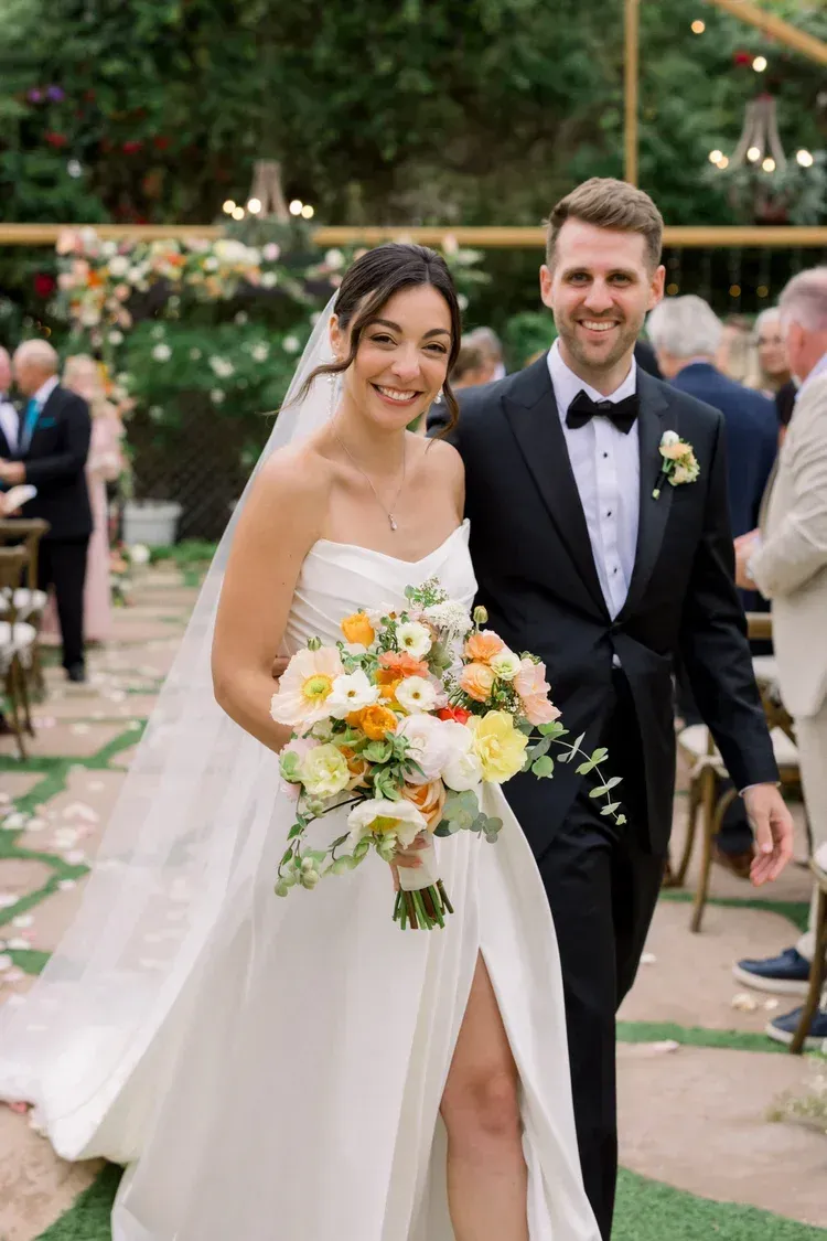 Bride and groom walking outdoors, bride holding a bouquet and smiling in a white gown, groom in a dark suit
