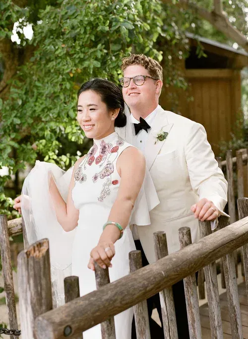 Two people in white formalwear leaning on a rustic wooden fence outdoors under trees