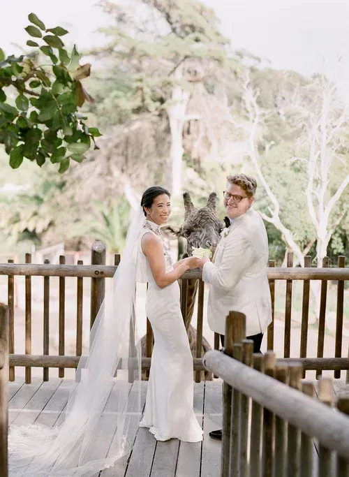 Couple in white wedding attire holding hands on a wooden deck with trees in the background