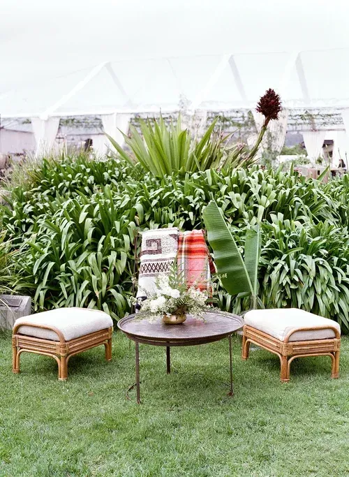 Outdoor lounge setup with wicker chairs, round table, and lush green plants under a white canopy.