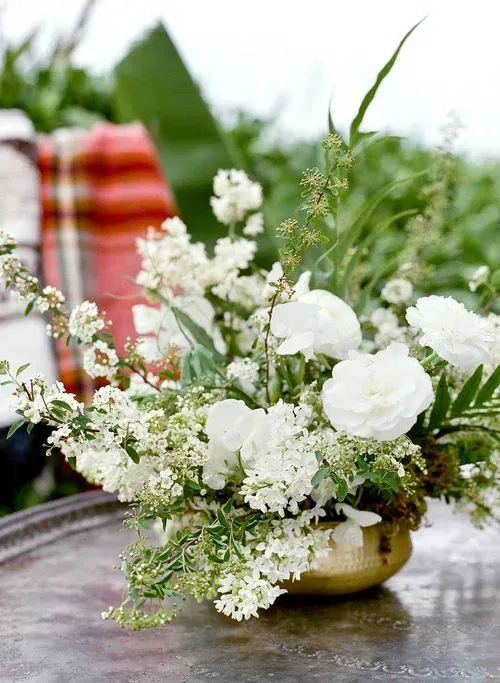White flowers in a gold vase on a rustic table with a blurred plaid chair in the background